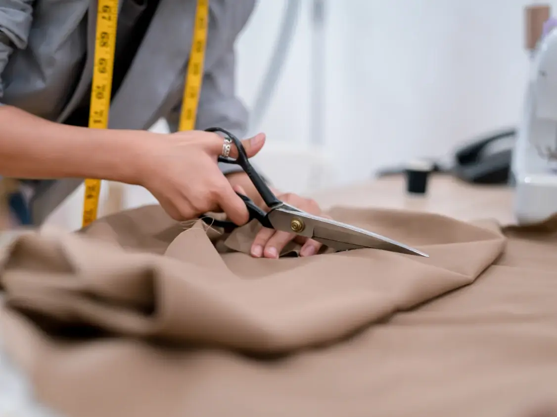 A fashion designer wearing a yellow measuring tape cuts tan or camel-colored fabric with large black-handled shears at a worktable