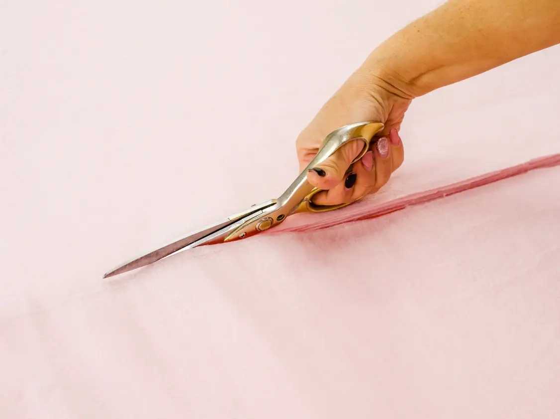 A hand with dark nail polish uses large gold-handled fabric scissors to cut pink fabric along a straight line