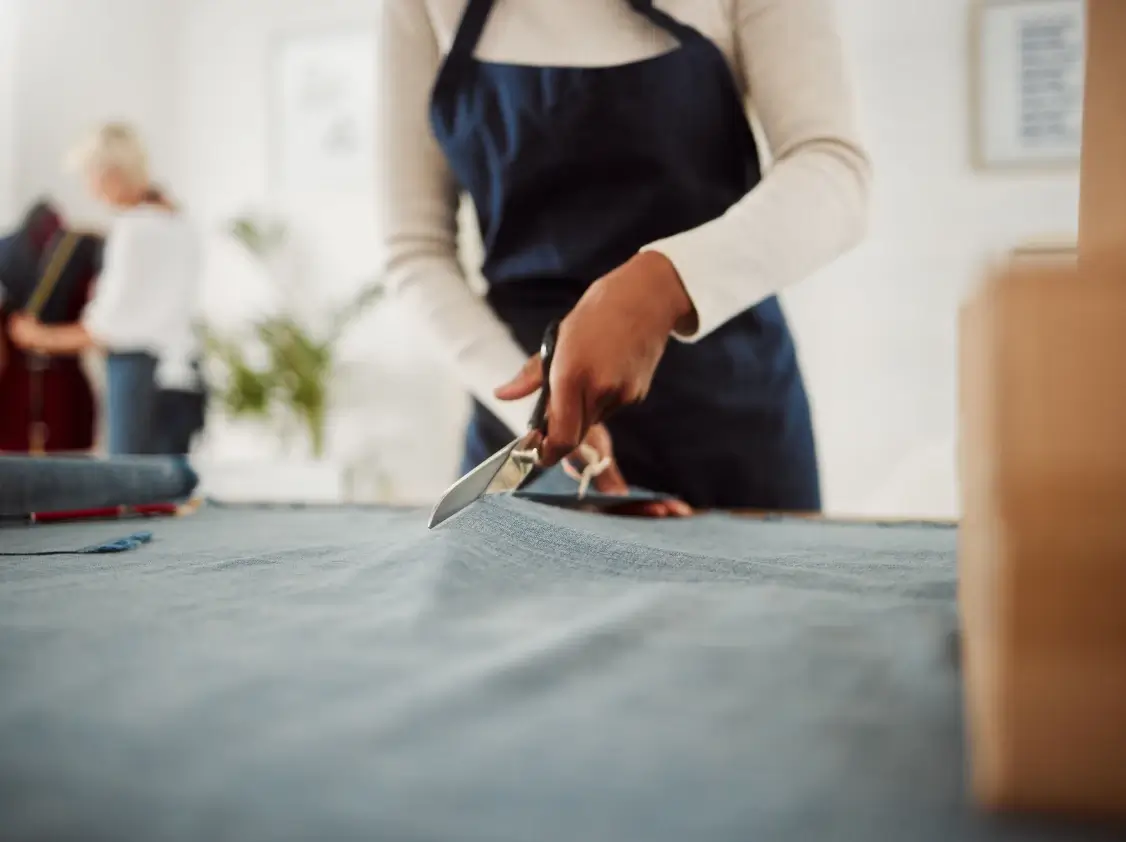 A person wearing a navy apron cuts blue-gray fabric with scissors at a cutting table in a bright sewing studio, with another person working in the background