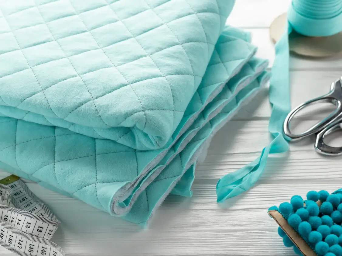 Folded mint green quilted fabric with diamond stitching pattern, surrounded by sewing notions including scissors, thread, bias tape, pom-pom trim, and a measuring tape on a white wood surface